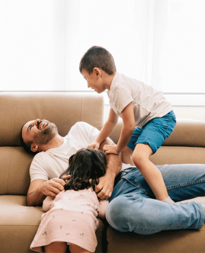 Father laughing with two young children during roughhousing play on a couch, showing joyful connection.