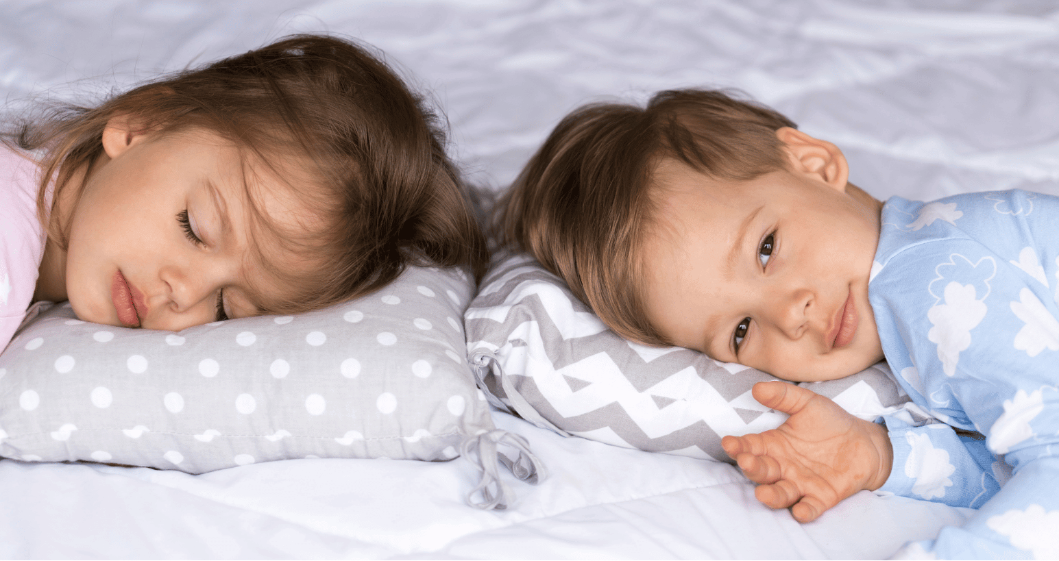 Twin toddlers lying in bed, one asleep and one awake.