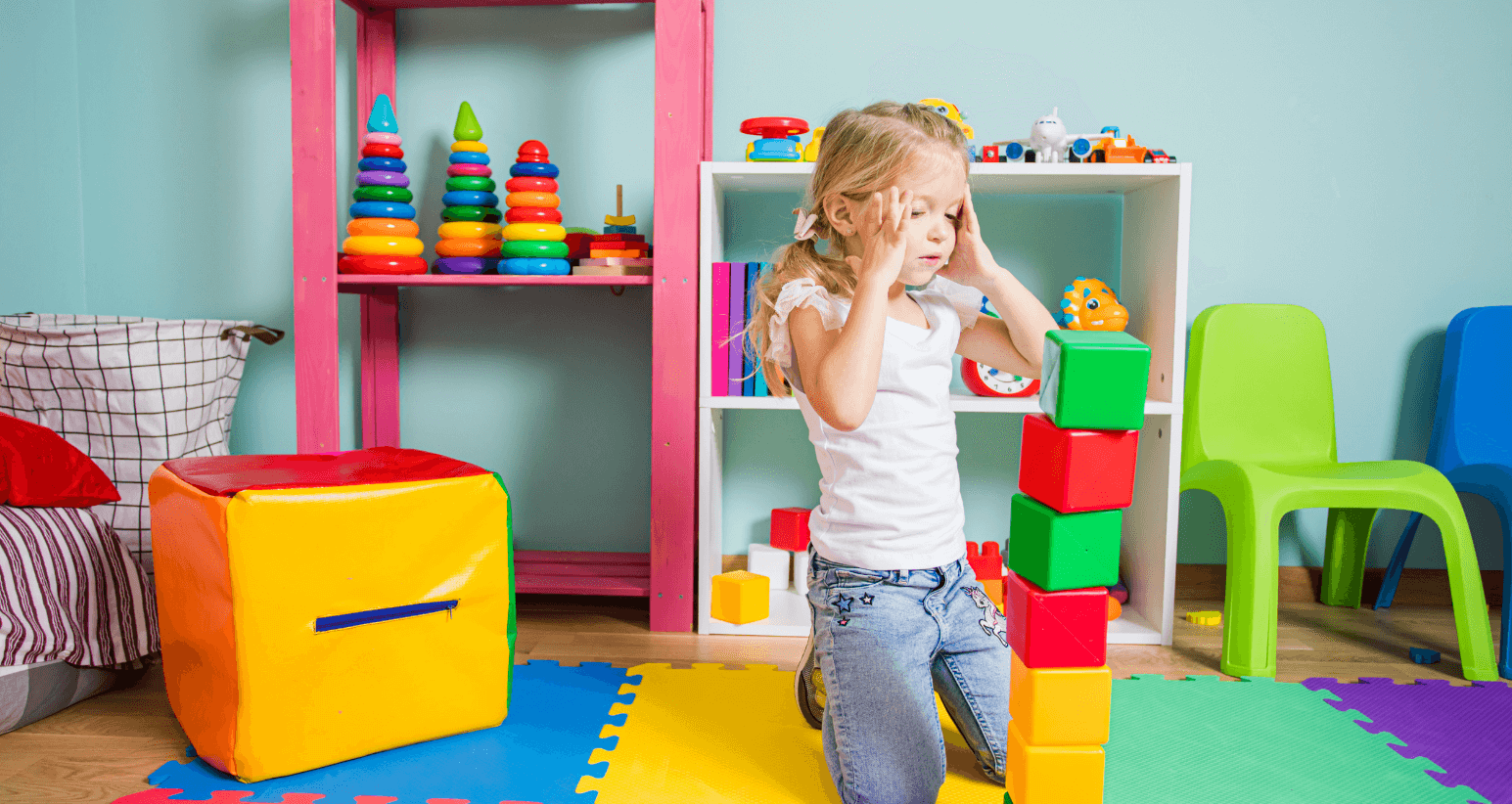 Frustrated young girl holding her head while playing with building blocks in a colorful playroom.