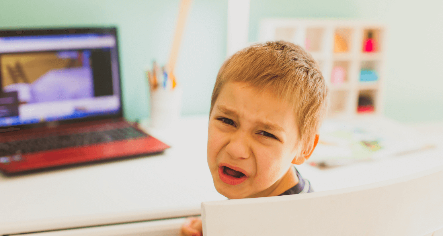 Upset young boy crying at his desk with a laptop in the background, expressing frustration and distress.