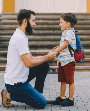 Father kneels to connect with young son carrying a backpack outdoors.