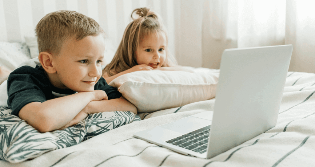 Young children smiling while watching a laptop on a bed.