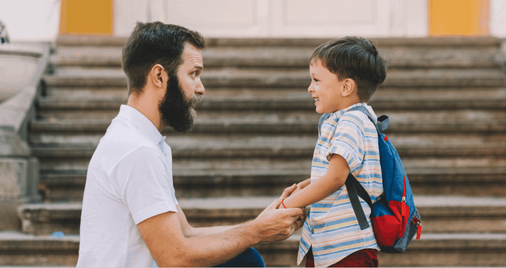 Father kneels to connect with young son carrying a backpack outdoors.