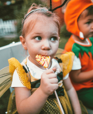 Child in Halloween costume eating a lollipop at a party.