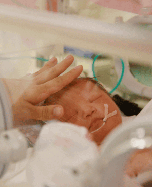 Newborn baby in NICU incubator with a caregiver’s hand gently resting on their head.