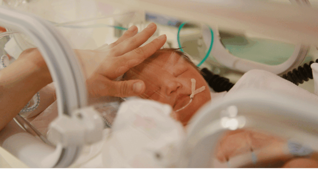 Newborn baby in NICU incubator with a caregiver’s hand gently resting on their head.