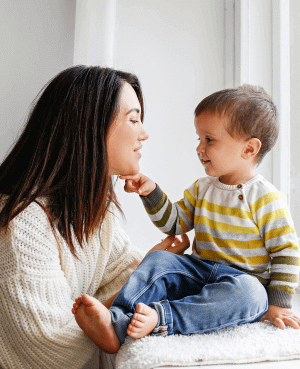 Parent and toddler smiling and touching hands while sitting near a window.
