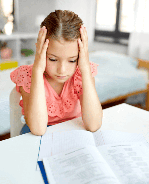 Child holding her head over homework, showing overwhelm from academic pressure.