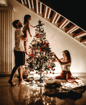 Parents and child decorating a lit Christmas tree together in a cozy home.