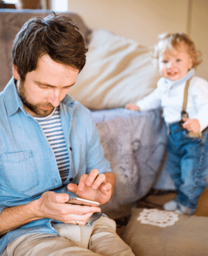 Dad looking at his phone while a toddler stands behind him, showing everyday distraction and screen time challenges.