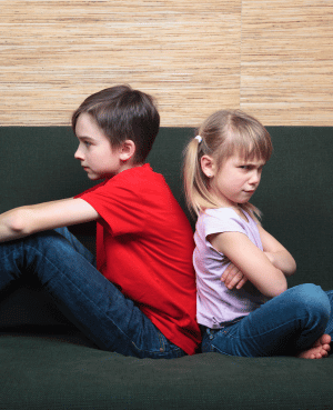 Two children sitting back to back on a couch, showing sibling conflict and fairness tension.