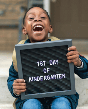 Smiling child holding a sign that says first day of kindergarten, showing kindergarten readiness excitement.