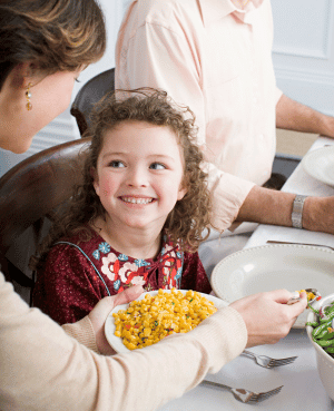 Child smiling at the dinner table while a parent serves food, representing positive mealtimes and confident eating.